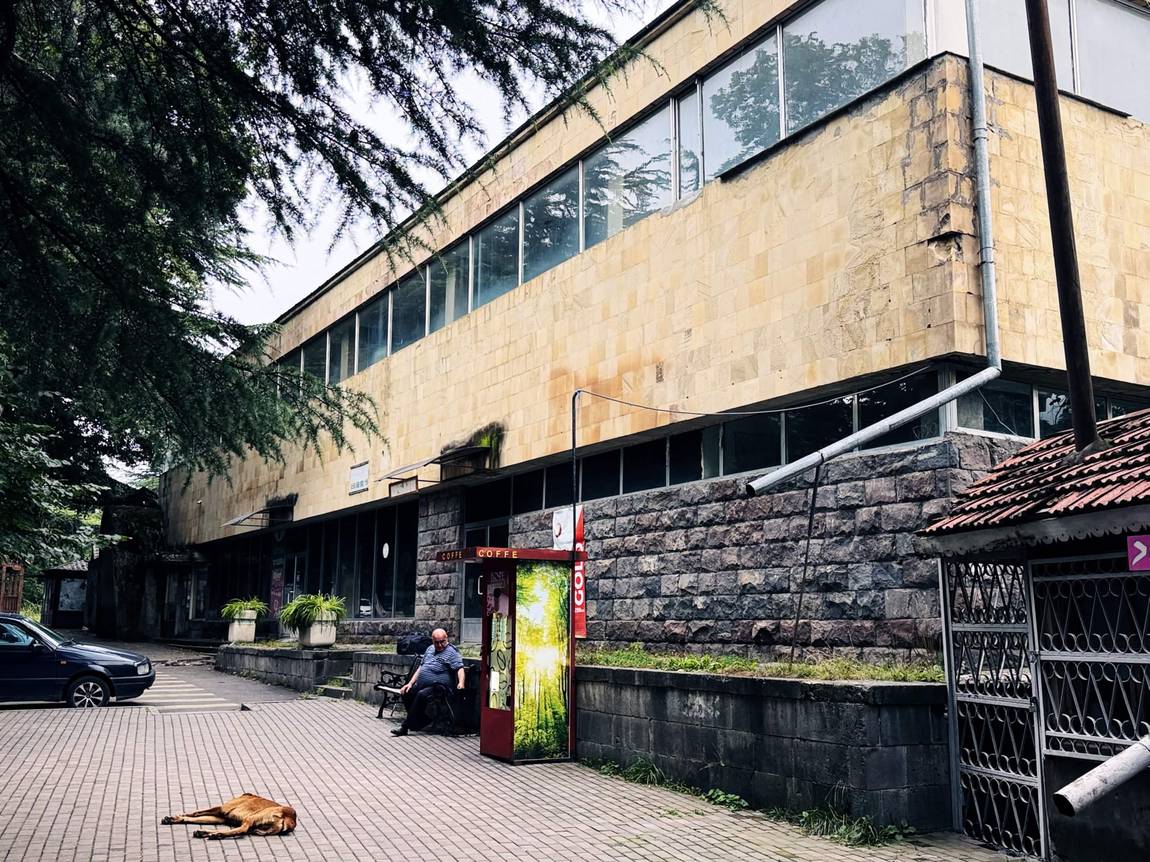 兩層樓的蘇聯時期商場，上層蜜黃色凝灰岩貼面配整條帶狀開窗，下層深色粗面石基座，門前有紅色咖啡亭、長椅上坐著一位男子、磚地上躺著一隻狗