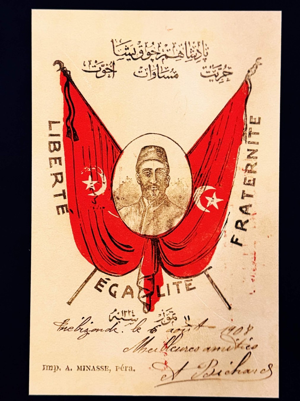 1908 年明信片，兩面交叉的紅色鄂圖曼星月旗中央為戴氈帽男子的橢圓肖像，兩側直排印有法文 LIBERTE 與 FRATERNITE，下方 EGALITE，頂端為鄂圖曼土耳其文口號，底部有手寫法文與印刷所名 Imp. A. MINASSE, Péra