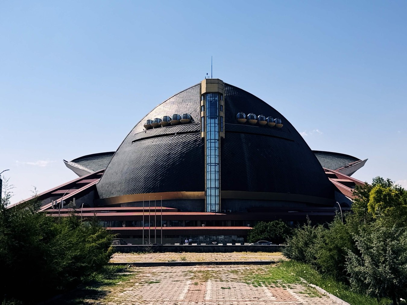 晚期蘇聯建築正面全景，深色金屬弧面屋頂如覆碗，兩側伸出翼狀懸挑，中央一道玻璃帷幕脊柱貫穿至頂，弧面上排列多個球形艙體，前方引道磚縫長草龜裂