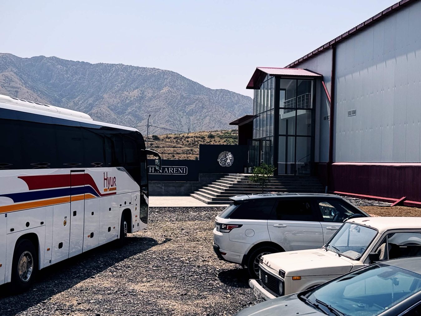Hin Areni 酒莊外的碎石停車場，Hyur Service 遊覽車停在門口，車身有紅藍橘三色條紋，背景是乾燥的褶皺山