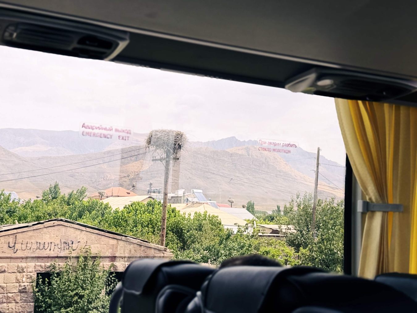 從遊覽車窗看出去，村莊電線桿頂端一個巨大的白鸛枝條巢，背景是乾燥的山與民宅鐵皮屋頂