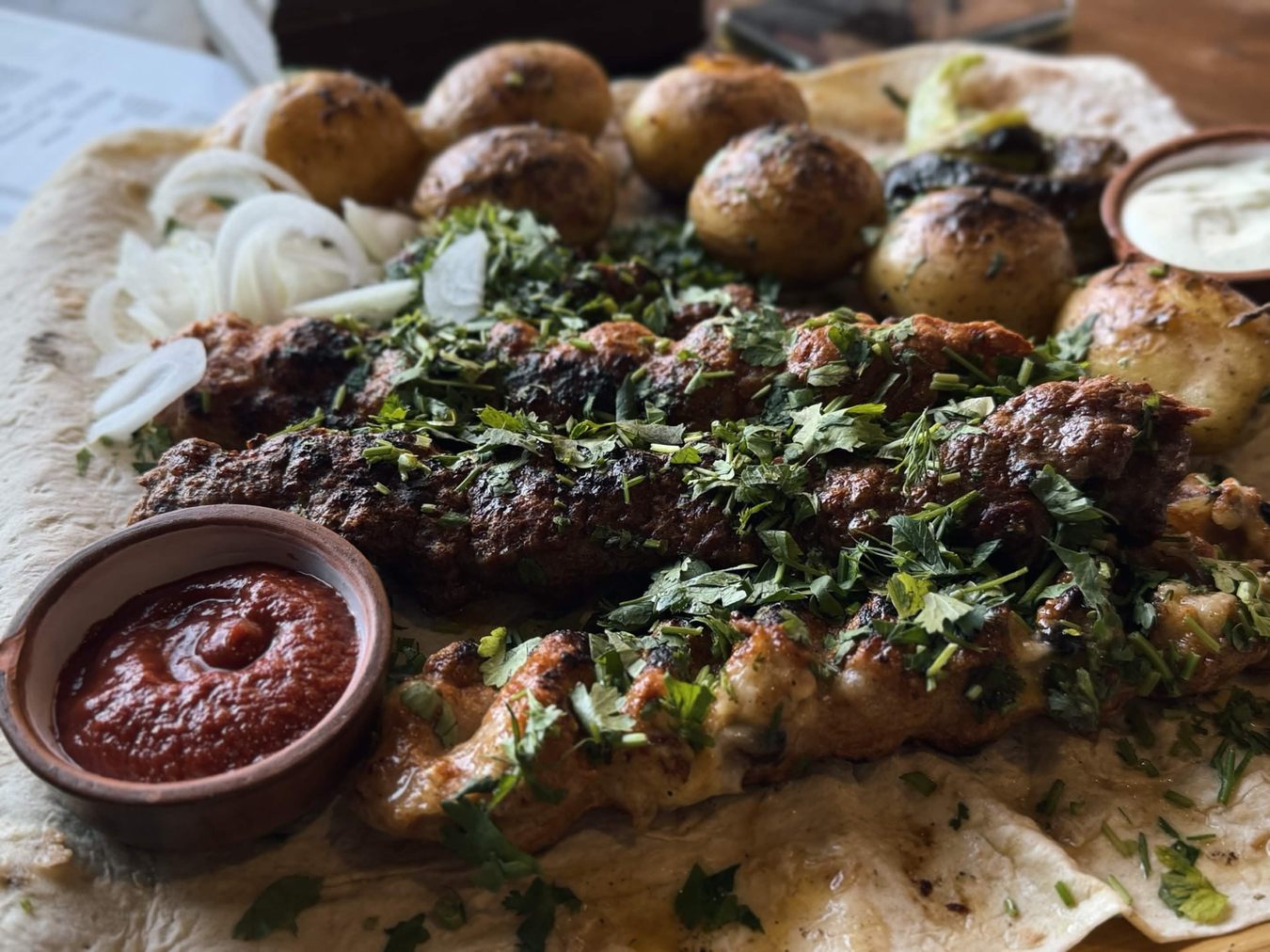 鋪在 lavash 上的烤肉拼盤：數支長條肉串、烤馬鈴薯、生洋蔥絲、番茄醬與優格醬陶碗