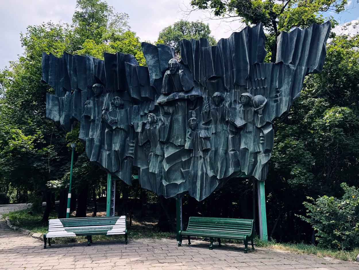 公園裡的大型青銅浮雕，架在細鋼柱上懸空，中央長袍人物周圍環繞多位手持書冊的年輕人