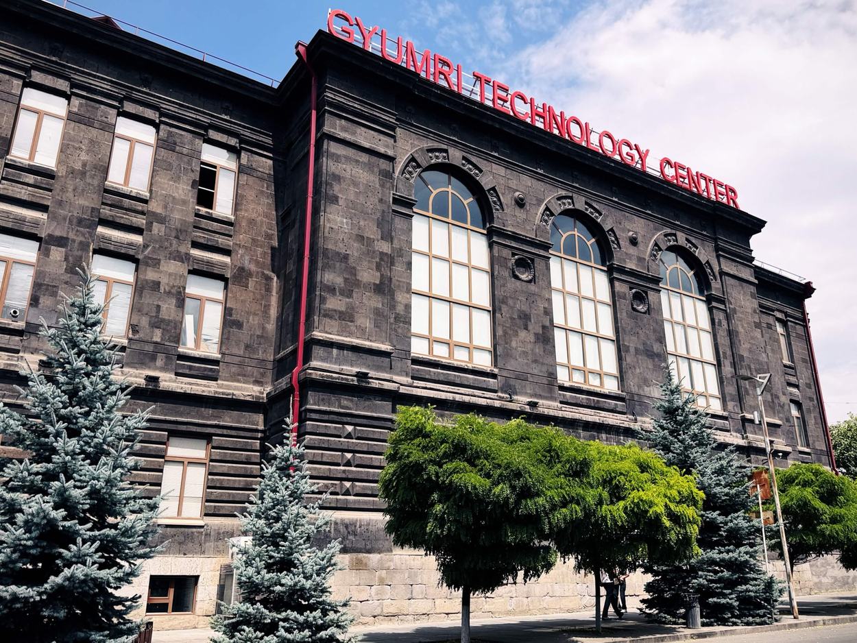 黑色凝灰岩的老建築，拱窗與壁柱，屋頂立著紅色 GYUMRI TECHNOLOGY CENTER 字樣招牌