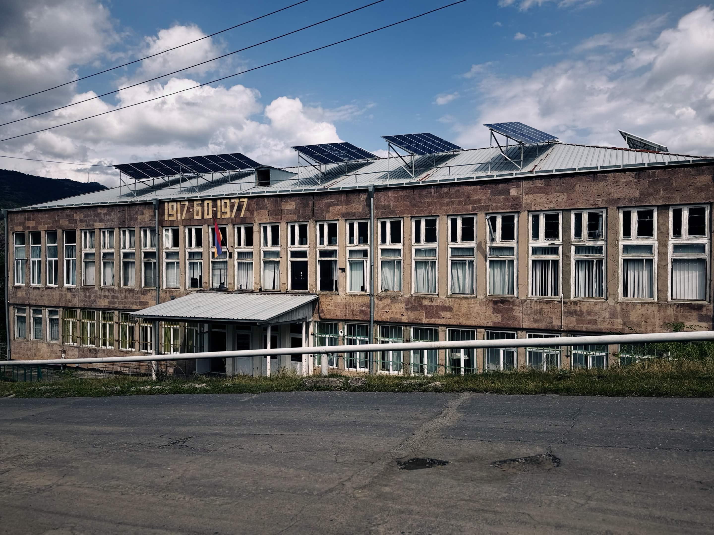 兩層石造校舍，正面掛著金色的 1917 60 1977 字樣，屋頂架著四片太陽能板，中央飄著亞美尼亞國旗